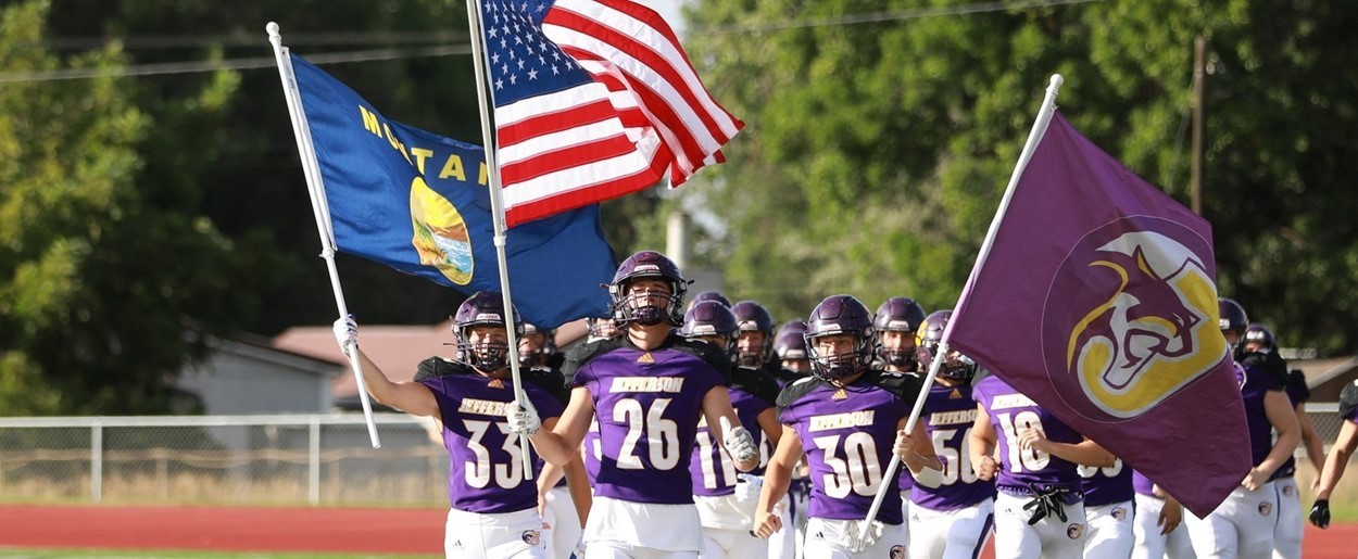 JHS football players carrying American, Montana, and JHS flags onto football field before game